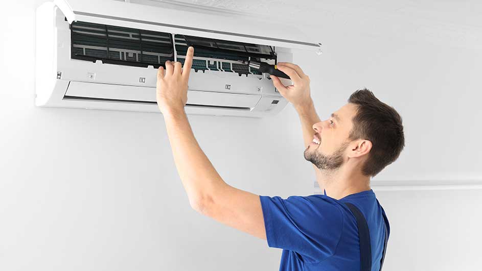 Un homme répare un climatiseur blanc fixé au mur blanc. Il utilise un outil noir.
