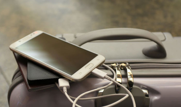Un smartphone blanc est posé sur une batterie externe noire, branché à un câble sur une valise grise.