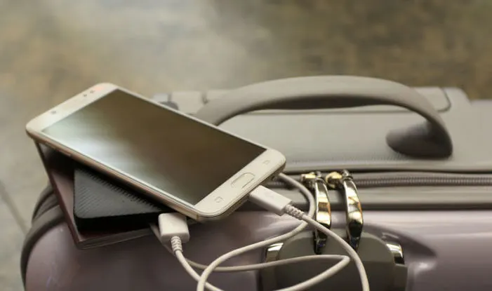 Un smartphone blanc est posé sur une batterie externe noire, branché à un câble sur une valise grise.