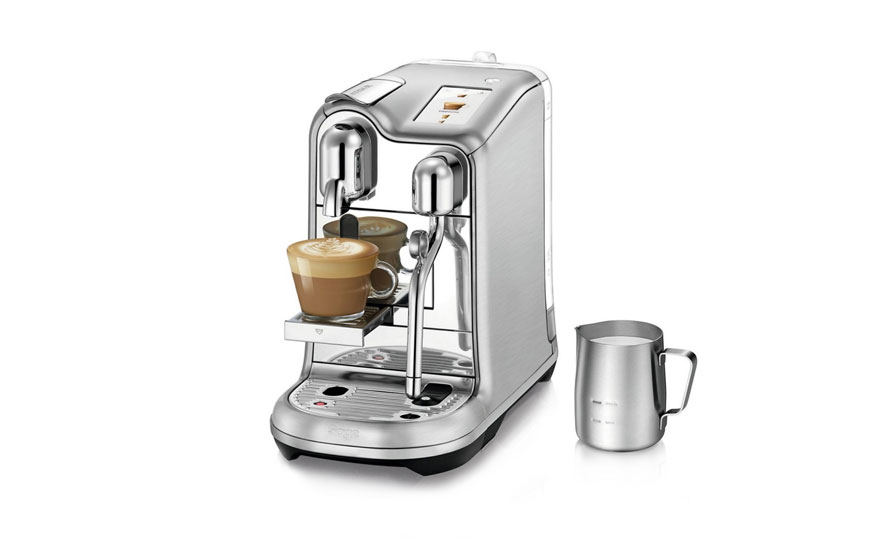 Een zilveren espressomachine met twee glazen kopjes koffie en een zilveren melkkan op een witte achtergrond.