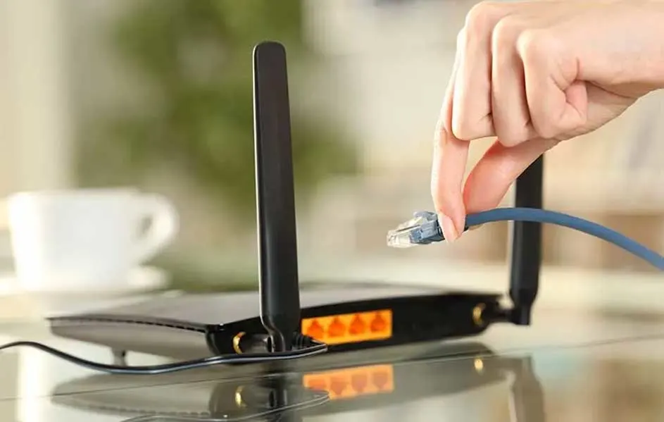 Een hand steekt een blauwe kabel in een zwarte router met twee antennes op een glanzend oppervlak.