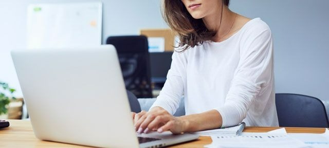 Een zilverkleurige laptop op een houten bureau met documenten. Op de achtergrond een bureaustoel en een wit bord.