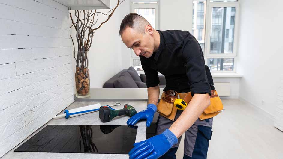 Un homme installe une plaque de cuisson noire. Il porte des gants bleus et une ceinture à outils en cuir marron.