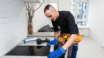 Un homme installe une plaque de cuisson noire. Il porte des gants bleus et une ceinture à outils en cuir marron.
