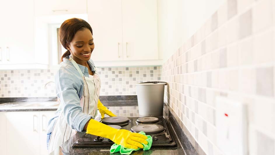 A kitchen scene shows a stovetop being cleaned with a green cloth and yellow gloves, featuring a bucket and tiled wall.