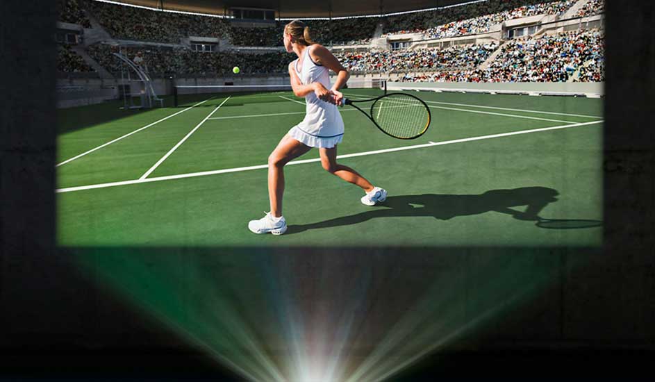 A tennis match is projected on a wall, showing a player in action on the court. A projector beams light to the screen. No brand is visible.