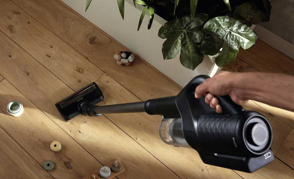 Une main tient un aspirateur noir nettoyant un plancher de bois clair. Des jouets en bois colorés sont dispersés sur le sol. Une plante verte est visible à l'arrière.