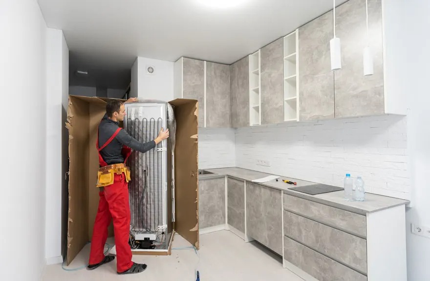 Un installateur en salopette rouge installe une nouvelle réfrigérateur dans une cuisine moderne avec des armoires grises. Marque : aucune visible.