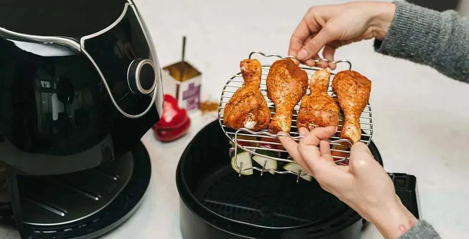Een zwarte airfryer staat naast kippenpoten op een rooster met groenten. Handen houden het rooster vast.