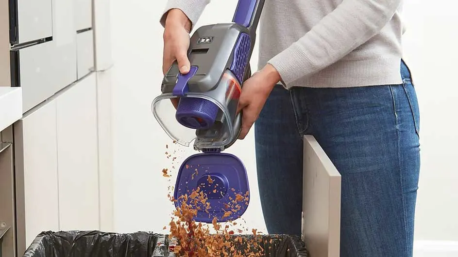 Une personne vide un aspirateur à main gris et violet au-dessus d'une poubelle noire. Des miettes brunes tombent. L'arrière-plan est blanc.