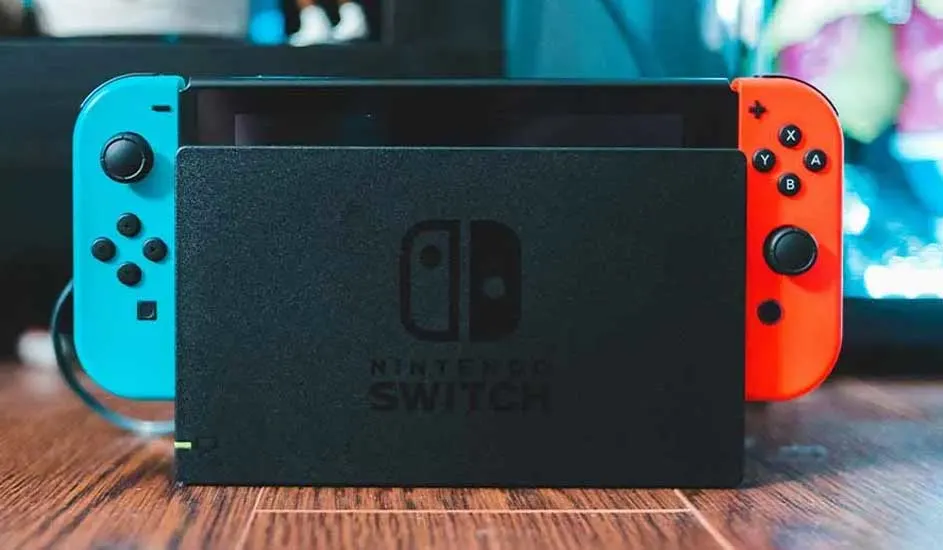 Une Nintendo Switch avec Joy-Con rouge et bleu est posée dans son dock noir, sur un sol en bois, éclairée par un arrière-plan coloré.