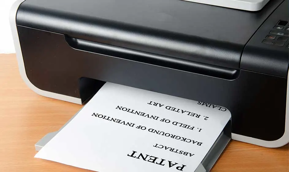 Une imprimante noire compacte fait sortir une feuille de papier. Le document présente des mots inversés et des lignes, posé sur un bureau en bois.