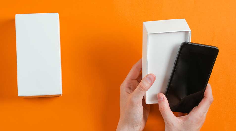 Twee witte dozen en een zwarte smartphone tegen een oranje achtergrond. Een hand houdt een open doos met een smartphone erin.