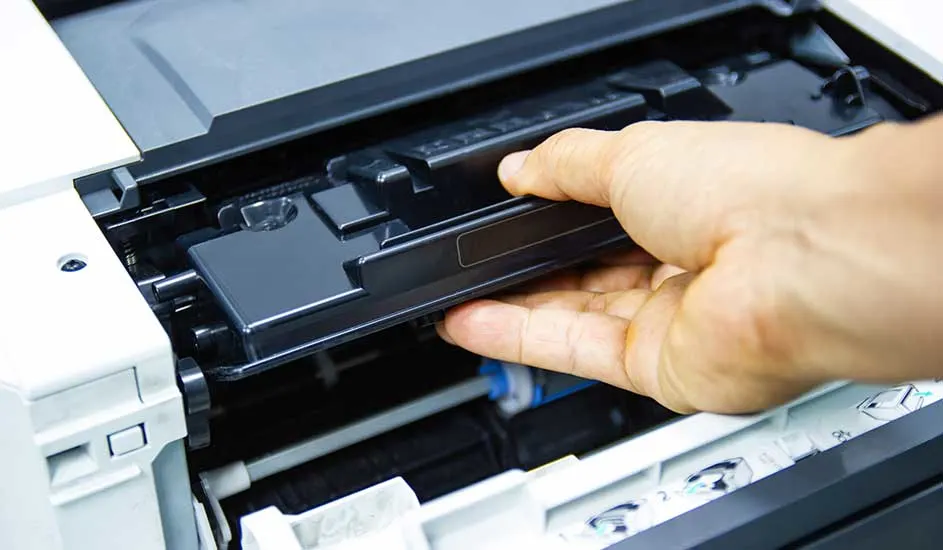 Een hand verwijdert een zwarte tonercartridge uit een witte en zwarte printer. Binnenkant zichtbaar.