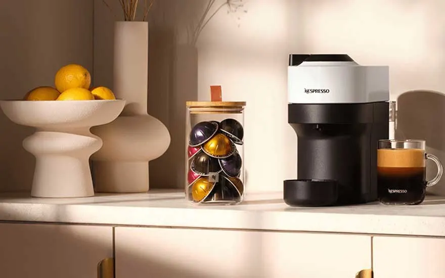 Witte Nespresso koffiemachine op marmeren aanrecht met glazen pot capsules, sinaasappels en verse espresso.