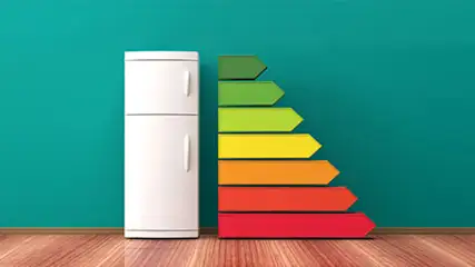 Witte koelkast naast energie-efficiëntielabel met pijlen in groen, geel, oranje en rood tegen een blauwe muur. Houten vloer.
