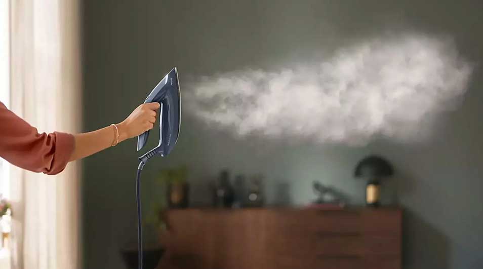Une main tient un défroisseur bleu qui émet de la vapeur. En arrière-plan, un mur vert et une commode en bois.
