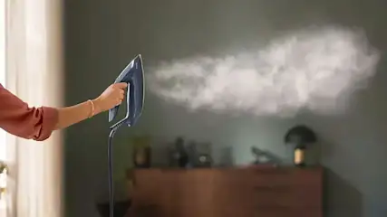 Une main tient un défroisseur bleu qui émet de la vapeur. En arrière-plan, un mur vert et une commode en bois.