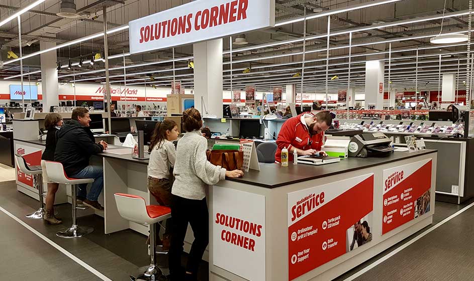 Een drukke 'Solutions Corner' in een grote winkel: klanten aan hoge balies, medewerkers helpen bij retouren, reparaties en advies.