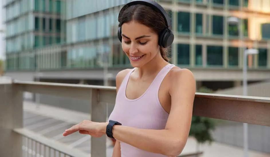 Een persoon met een roze top en zwarte hoofdtelefoon kijkt naar een zwarte smartwatch buiten, met een gebouw op de achtergrond.