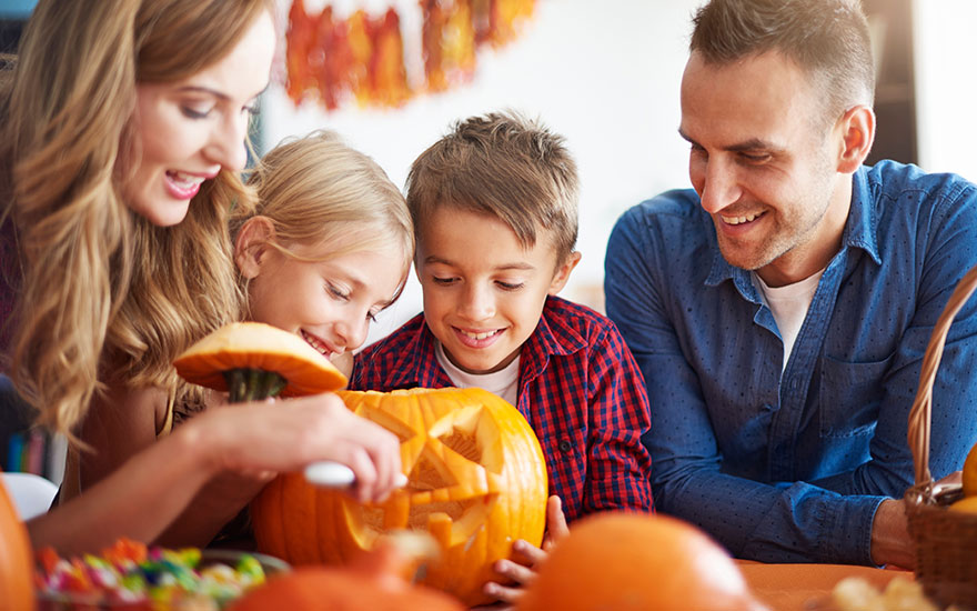 Une famille sculpte une citrouille orange d'Halloween avec des outils sur une table, entourée d'autres citrouilles et décorations.