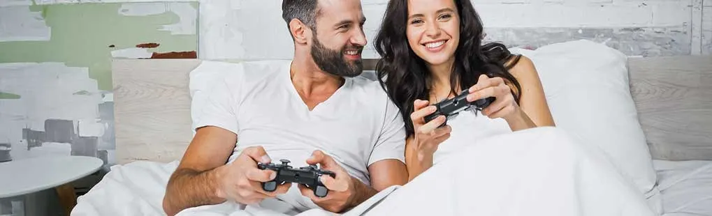 Een lachend koppel ligt samen in bed in witte lakens, ieder met een gamecontroller, ontspannen spelend in een lichte slaapkamer
