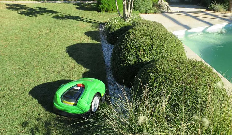 Een groene Ambrogio robotmaaier rijdt over het gras naast struiken en een zwembad in de zon.