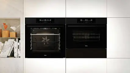 Twee zwarte ovens ingebouwd in witte kasten. Links een plank met boeken en decoratieve objecten.