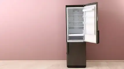 Een koelkast met open deur staat tegen een roze muur op een houten vloer. Het interieur is wit met glazen planken.