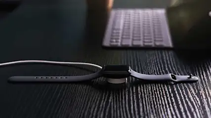 Une montre noire est en charge sur un bureau en bois sombre, avec un clavier flou à l'arrière-plan.