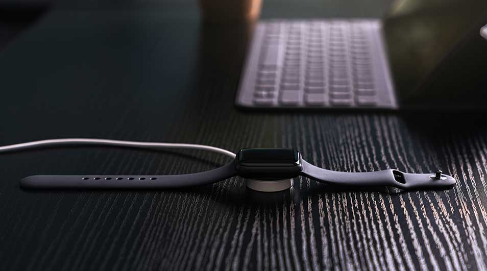 Zwarte smartwatch aan het opladen op een houten bureau, met een wit toetsenbord op de achtergrond.