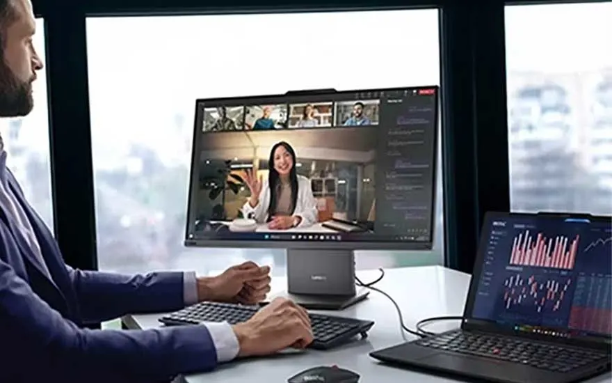 Een man zit aan zijn bureau met een groot scherm voor videogesprek en een laptop waarop rode en blauwe financiële grafieken staan.