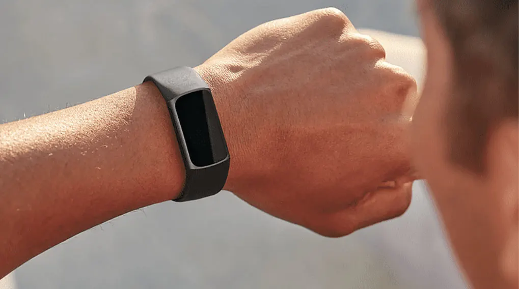 Poignet avec bracelet de suivi d'activité gris et écran noir, sur fond flou.