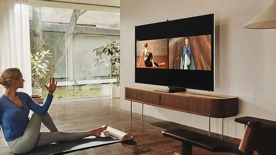Woonkamer met tv op dressoir, yoga mat op de vloer. Buiten zichtbaar door groot raam met gordijnen.