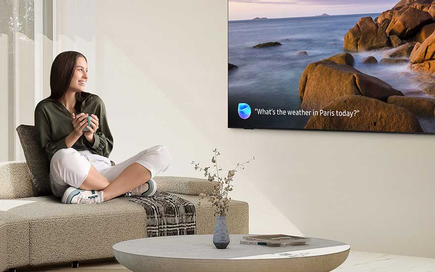 Une jeune femme détendue sur le canapé, tasse à la main, regarde une télévision moderne avec assistant vocal.