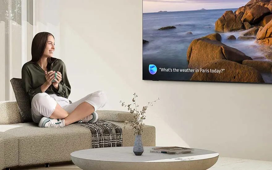 Een jonge vrouw zit ontspannen op de bank met een beker, glimlachend en kijkend naar een moderne tv met spraakassistent.