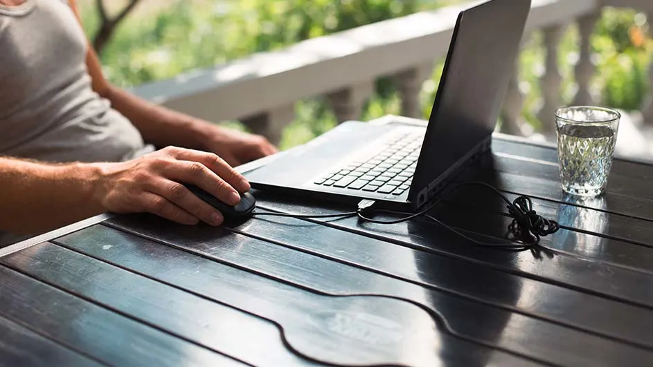 Een laptop en een muis op een houten tafel, met een glas water ernaast. Buiten is er groen te zien.
