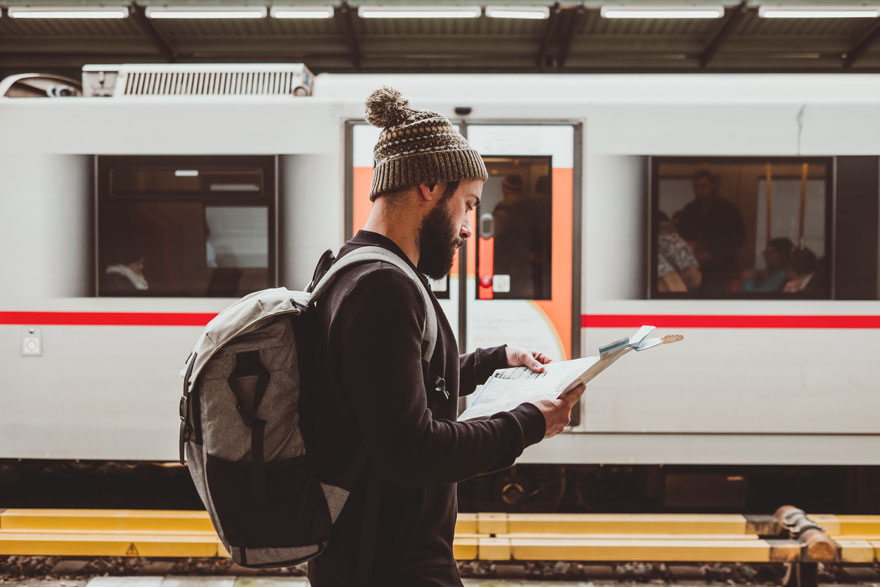 Un homme avec un sac à dos regarde une carte devant un train blanc avec une bande rouge sur un quai de gare.