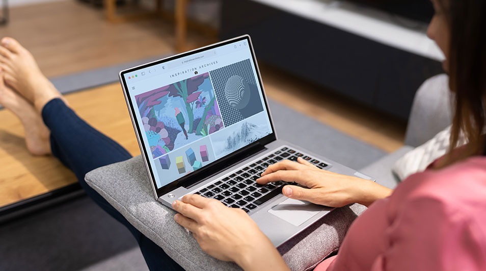 A laptop rests on a gray cushion, displaying a website with colorful images. Hands type on the keyboard. A wooden surface is nearby.