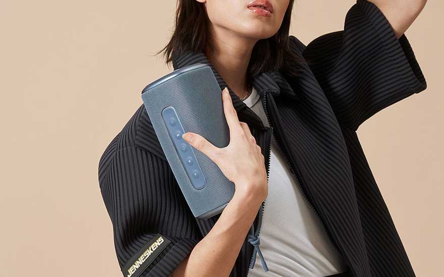 Haut-parleur cylindrique gris avec boutons bleus, veste rayée noire, fond beige.