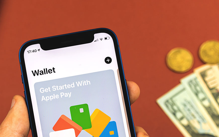 Gros plan d'un iPhone affichant l'application Wallet, avec des billets et des pièces en arrière-plan sur une surface rouge.