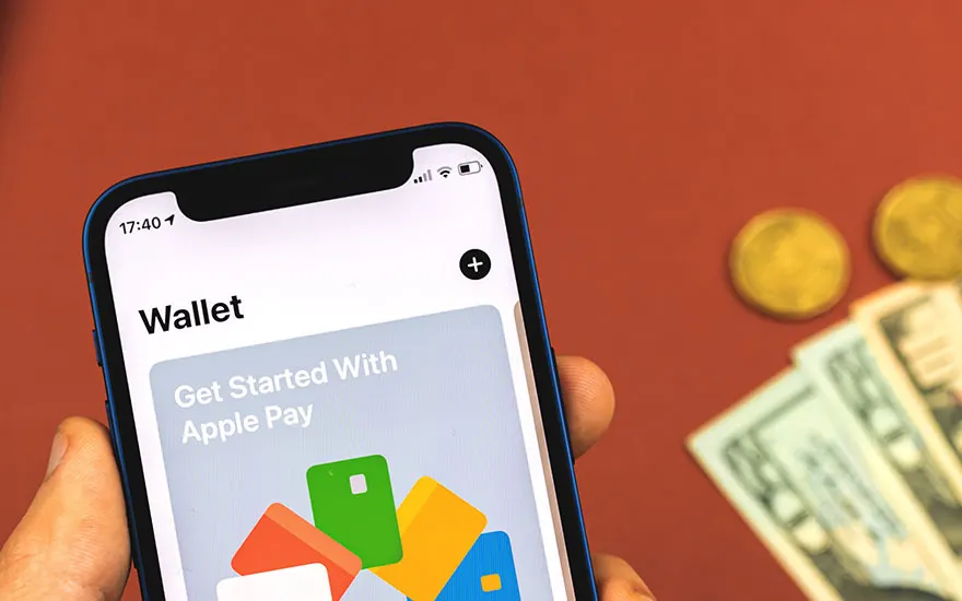 Gros plan d'un iPhone affichant l'application Wallet, avec des billets et des pièces en arrière-plan sur une surface rouge.