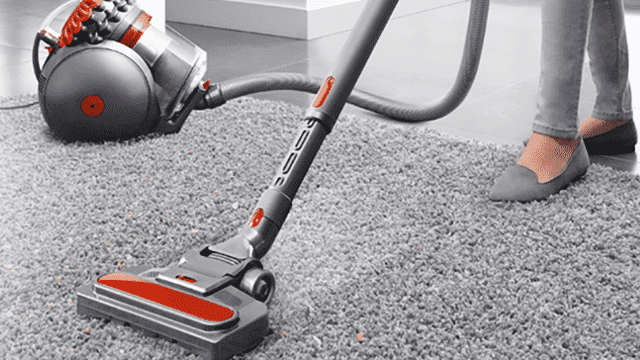 Un aspirateur gris et rouge est sur un tapis gris. Le tuyau est connecté à un réservoir rond.