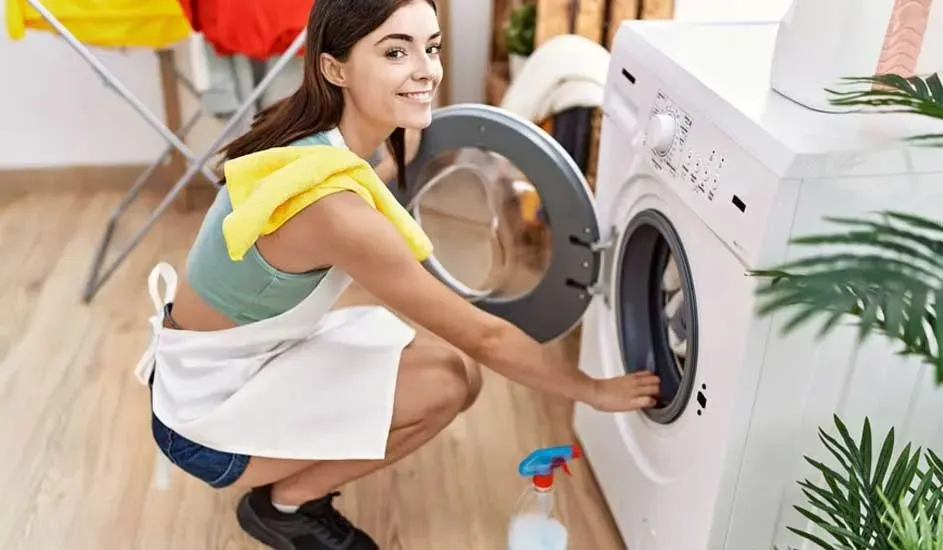 Een vrouw in sportkleding en schort laadt wasgoed in een witte wasmachine, merk niet zichtbaar, met schoonmaakmiddel op de vloer.
