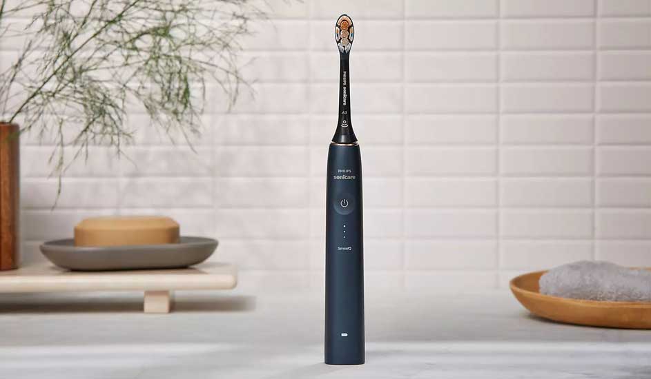 Une brosse à dents électrique bleue se dresse sur un comptoir de salle de bain carrelé, avec du savon, une serviette et des plantes.