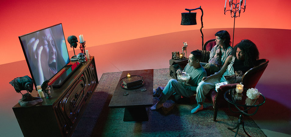 Salon avec téléviseur affichant un visage effrayé, tables basses, lampes, canapé et fauteuil sur un tapis à motifs.