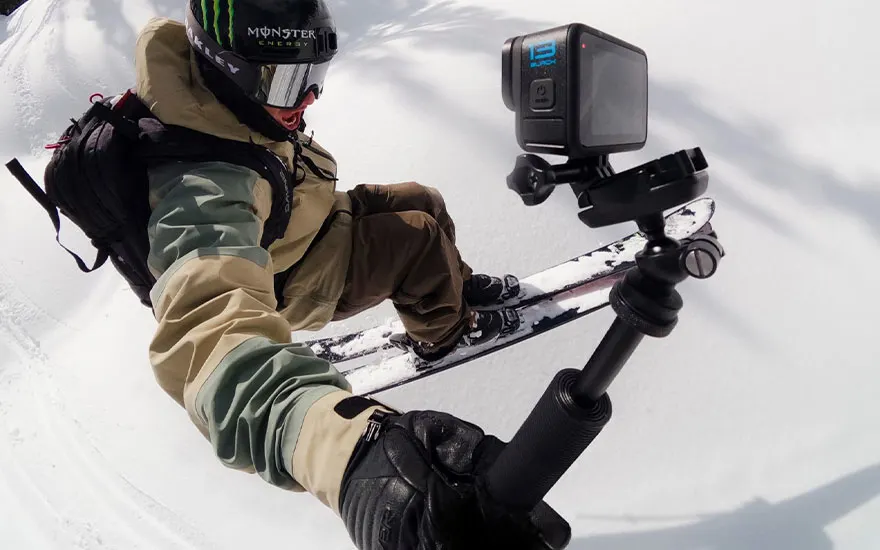 A snowboarder on skis holds a camera pole. Snow is in the background.