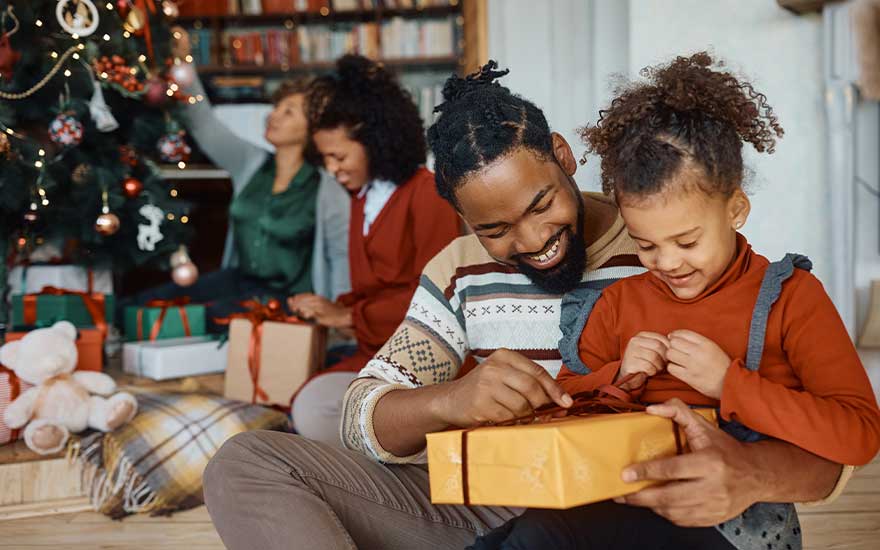 Un père et sa fille sont assis près du sapin de Noël, ouvrant joyeusement un cadeau jaune pendant que la famille décore derrière eux.