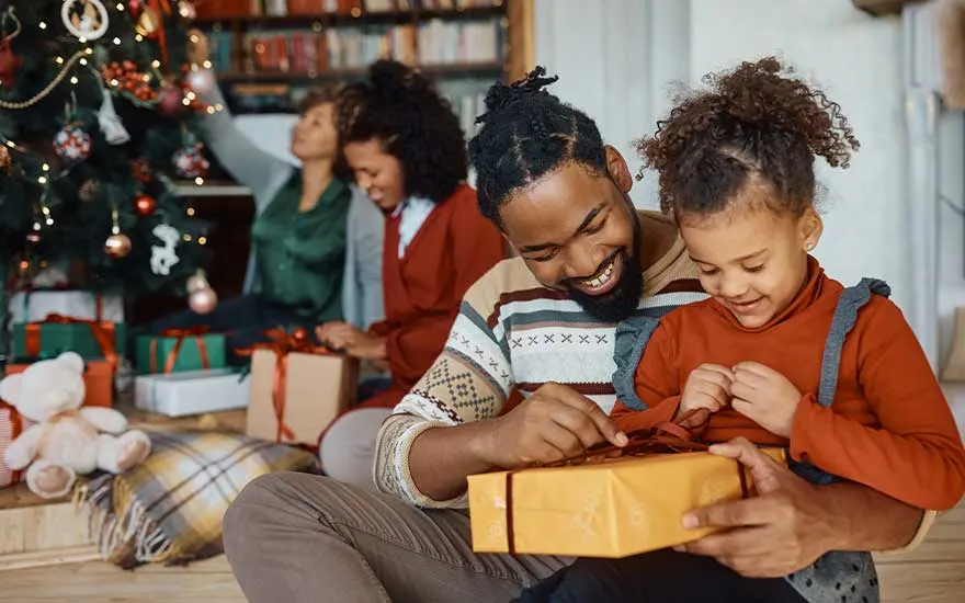 Un père et sa fille sont assis près du sapin de Noël, ouvrant joyeusement un cadeau jaune pendant que la famille décore derrière eux.