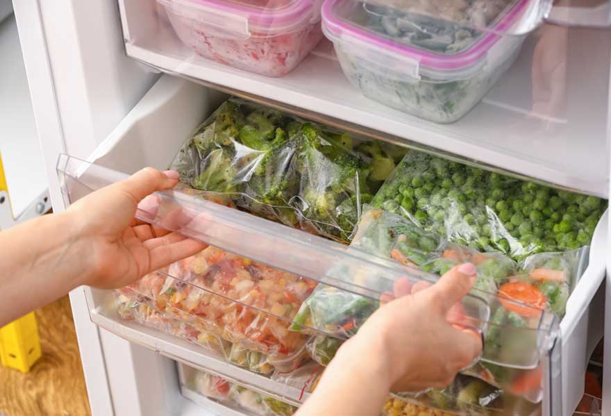 Een open witte vriezer met heldere plastic bakken vol ingevroren groenten, zoals broccoli, erwten en wortelen.